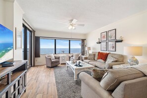 A spacious living room with a large window offering a view of the sea, a ceiling fan, and a fireplace with framed botanical prints. - Four Seasons 702E by Liquid Life (Orange Beach)