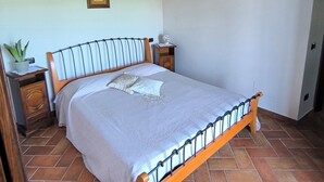Un dormitorio con cabecera de cama de madera, colcha blanca, mesita de noche con un jarrón y una planta en maceta.
