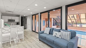 Un interior moderno con un sofá gris, sillas blancas, una zona de cocina y vistas a una piscina a través de amplios ventanales.