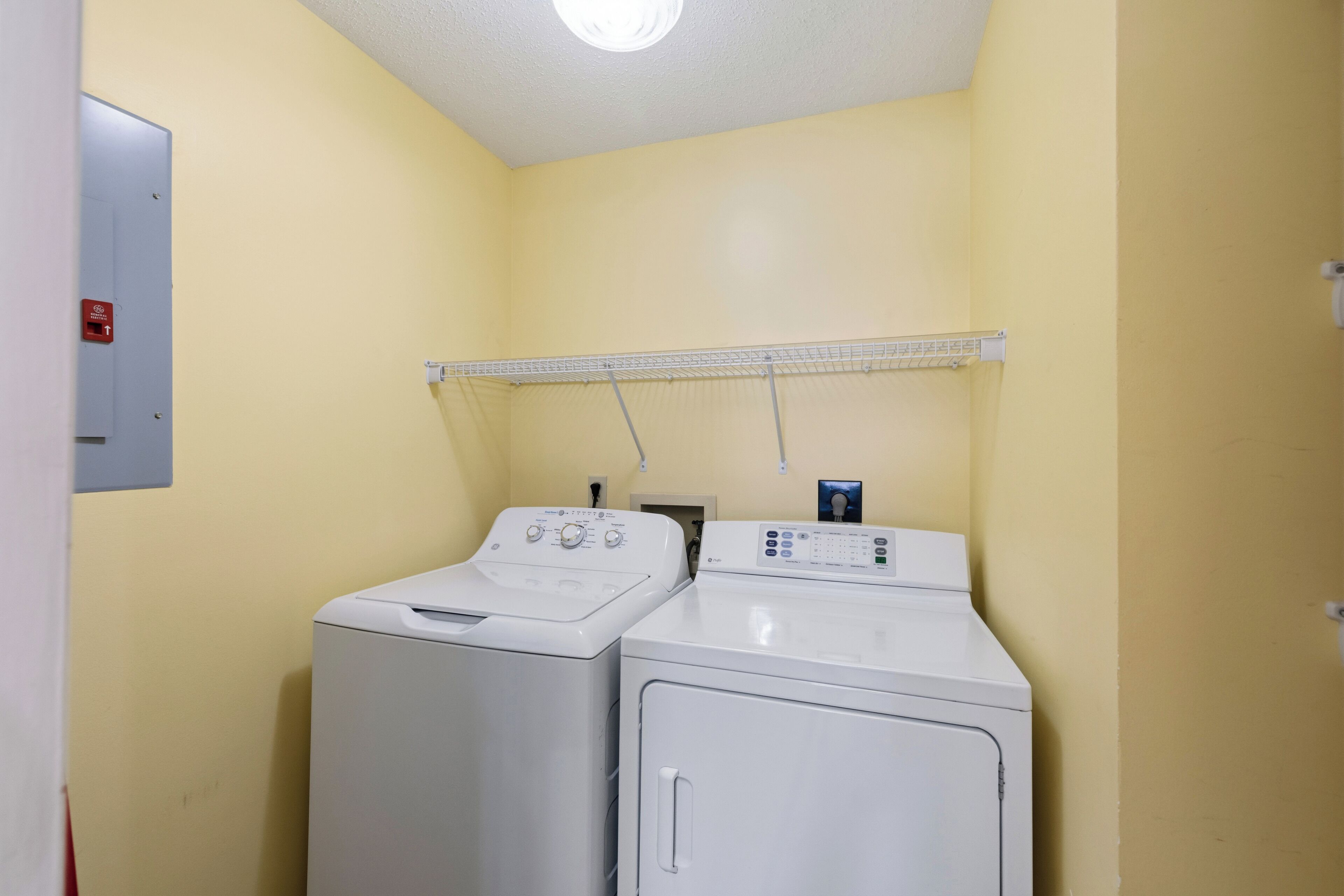 A laundry room with two front-loading washing machines and a dryer, a metal shelf above the machines, and a fire alarm on the wall.