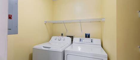 A laundry room with two front-loading washing machines and a dryer, a metal shelf above the machines, and a fire alarm on the wall.