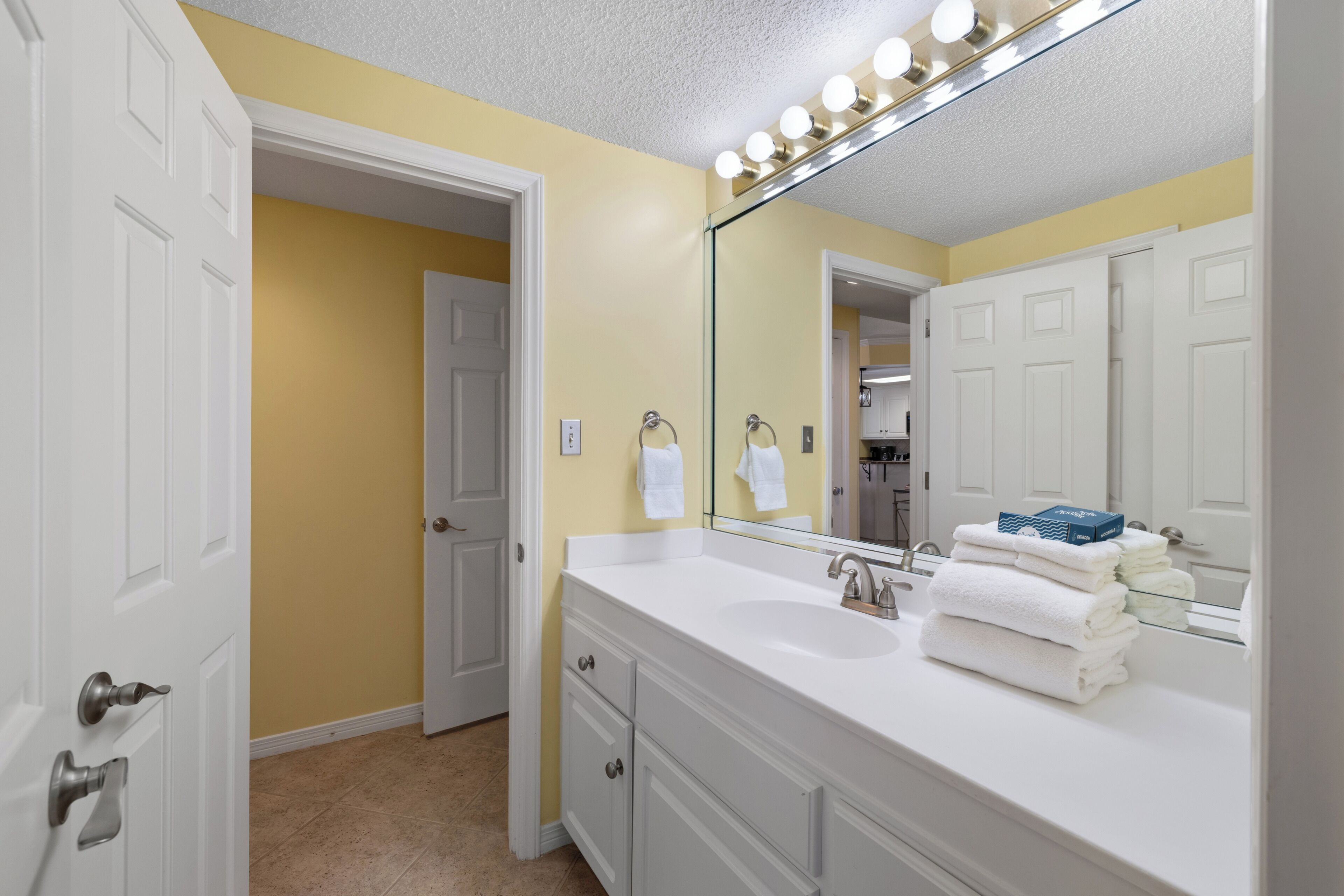 Une salle de bain avec un meuble-lavabo blanc, un grand miroir et un évier. Des serviettes sont pliées et posées sur le meuble-lavabo.