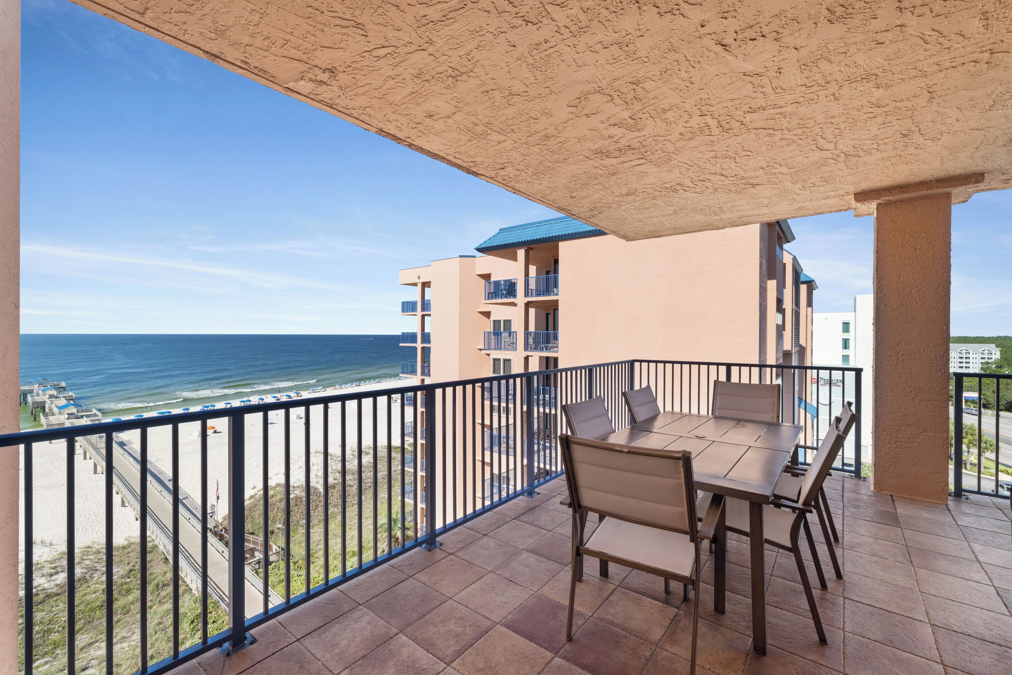 A balcony with a view of the beach and ocean, a table with chairs, and a building with balconies in the background.