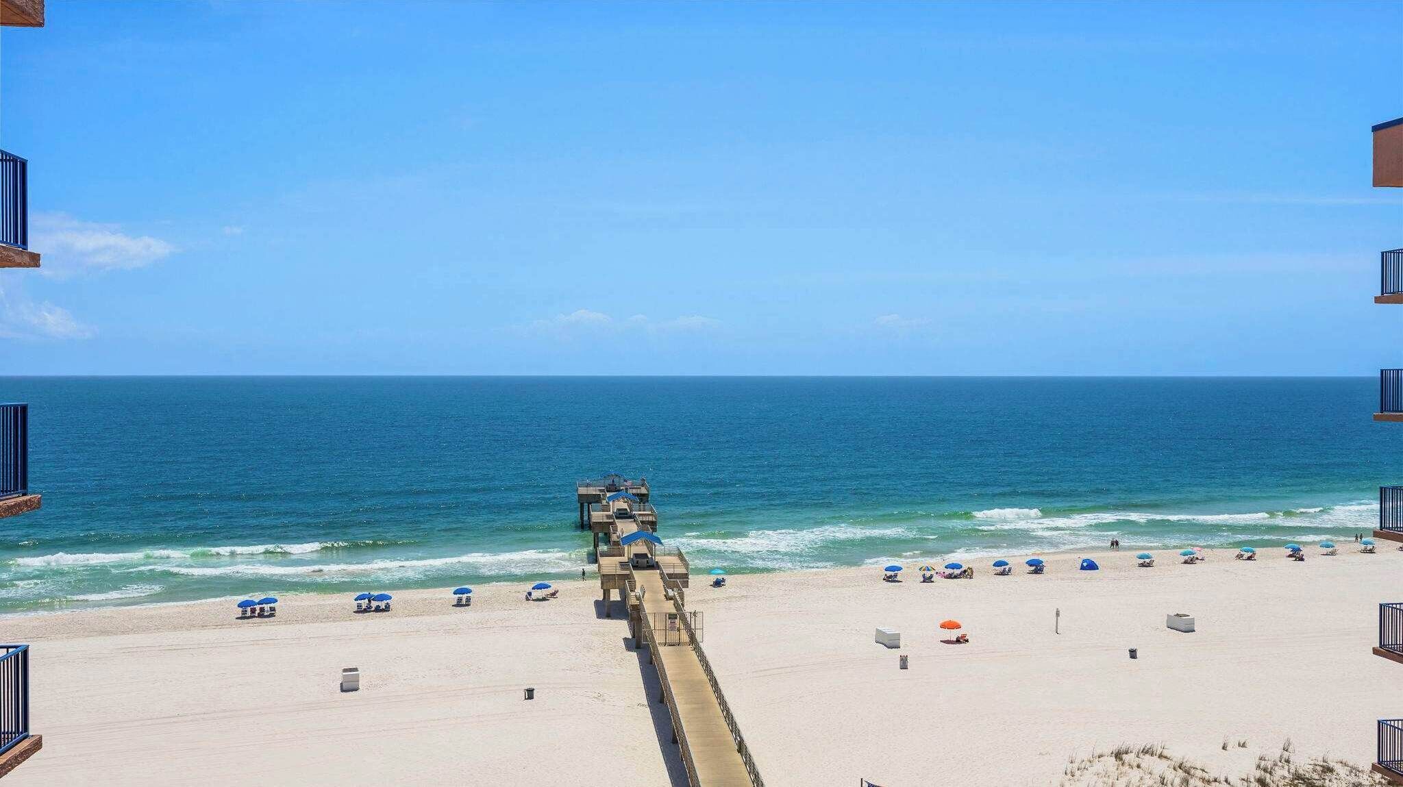 Una playa con un muelle que se adentra en el océano, sombrillas de playa y un cielo despejado.