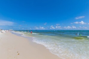 Ein Strand mit klarem Wasser, ein Pier in der Ferne und Menschen im Wasser.