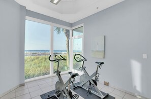A room with two stationary bikes, large windows with a view of the beach, and a painting on the wall. - Lighthouse 1505 by Liquid Life (Gulf Shores)