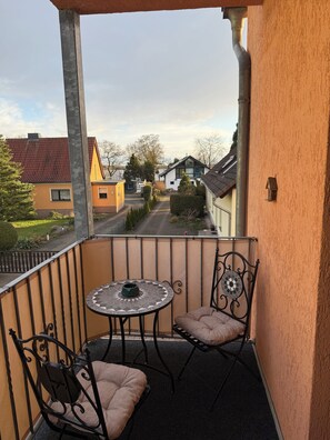 Ein Balkon mit rundem Tisch und zwei Stühlen, Blick auf eine Wohnstraße und ein Haus mit rotem Dach.
