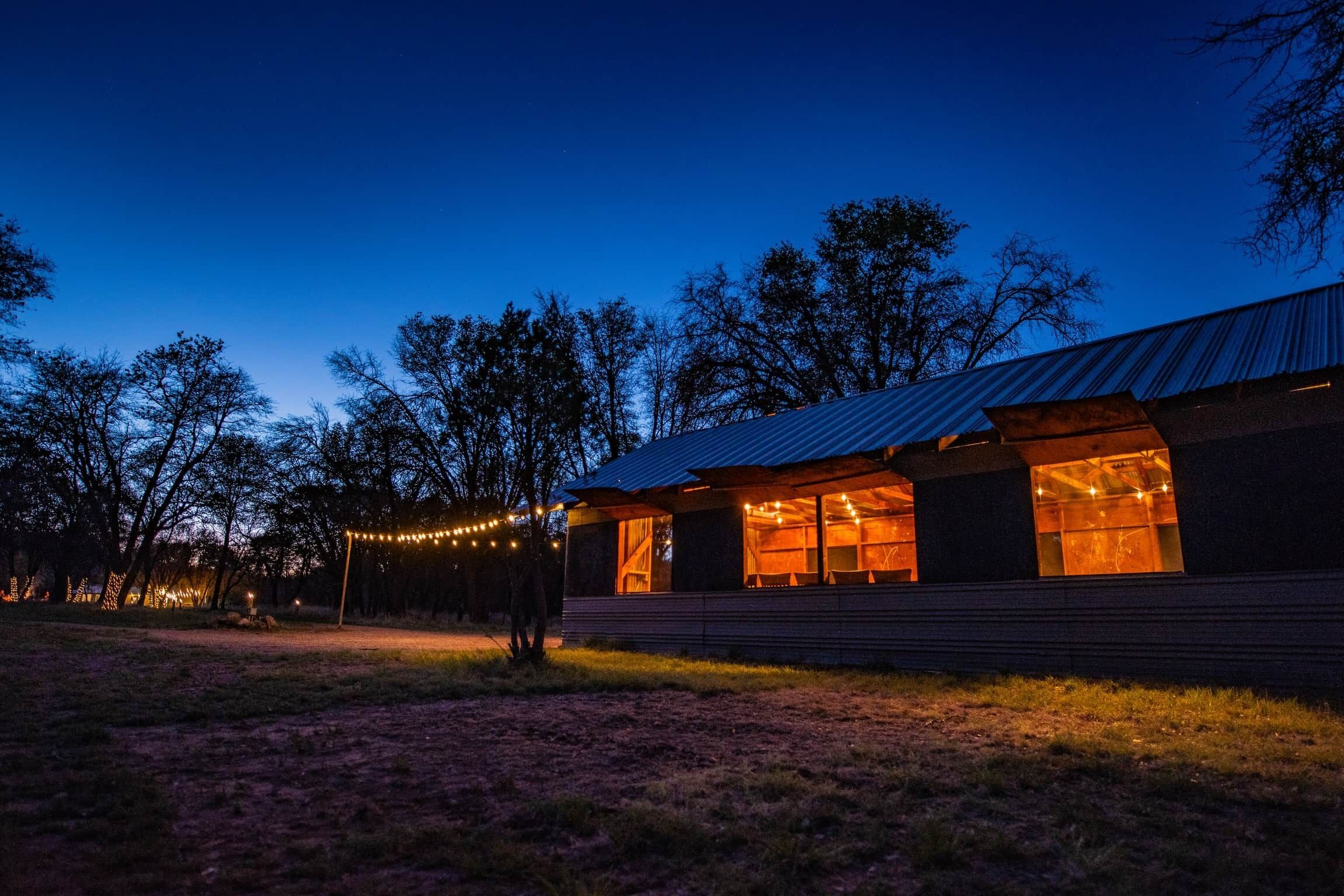Un bâtiment moderne doté de grandes fenêtres et d’un toit en métal, entouré d’arbres et illuminé de guirlandes lumineuses.