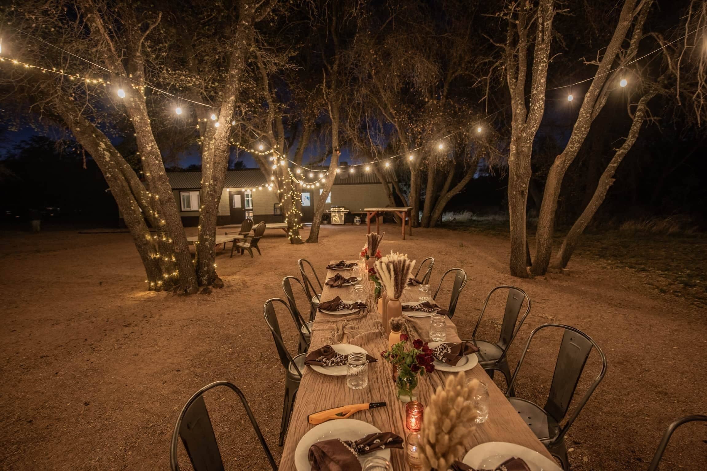 Un espacio exterior para comer con una larga mesa dispuesta para una comida, rodeado de árboles y luces colgantes.