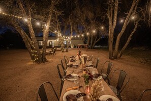 An outdoor dining area with a long table set for a meal, surrounded by trees and string lights.