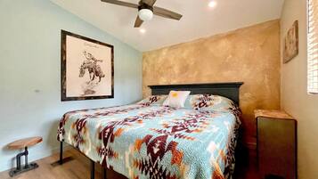 Une chambre à coucher avec un lit, un ventilateur de plafond, un tableau encadré au mur et une petite table.