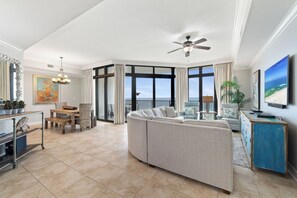 A spacious living area with a large sofa, dining table, and chairs, a ceiling fan, and a view of the ocean through large windows. - The Oasis at Orange Beach 710 by Liquid Life (Orange Beach)