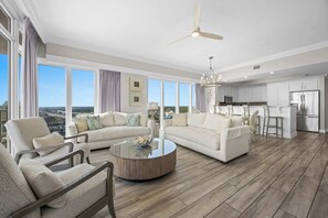 Ein geräumiges Wohnzimmer mit großem Fenster, Deckenventilator und moderner Küche im Hintergrund.