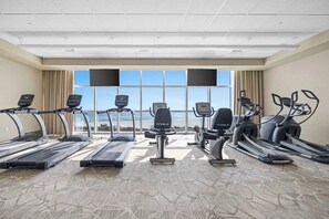 Ein Fitnessstudio mit Laufbändern, Crosstrainern und einem großen Fenster mit Blick auf den Strand.