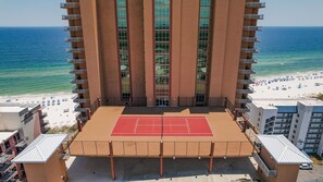 Un edificio de gran altura con una cancha de tenis en la planta baja, con vista a una playa con sombrillas y gente.