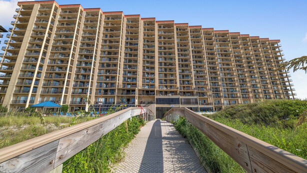 Un hotel de diverses plantes amb una entrada central, un camí de fusta que hi mena i un cel blau clar.