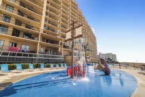 Una fuente de agua con estructura de cruz frente a un hotel de varios pisos con balcones y una zona de piscina.