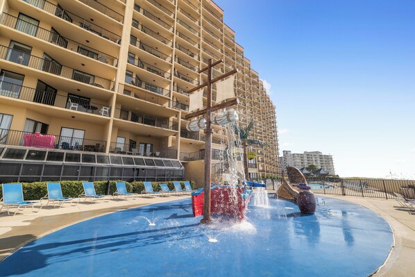 A water fountain with a cross structure in front of a multi-story hotel with balconies and a pool area. - Phoenix V 505 by Liquid Life (Orange Beach)
