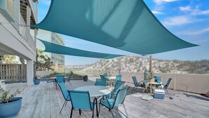 Una terraza de madera con mobiliario exterior, incluyendo una mesa y sillas, bajo una vela de sombra grande.