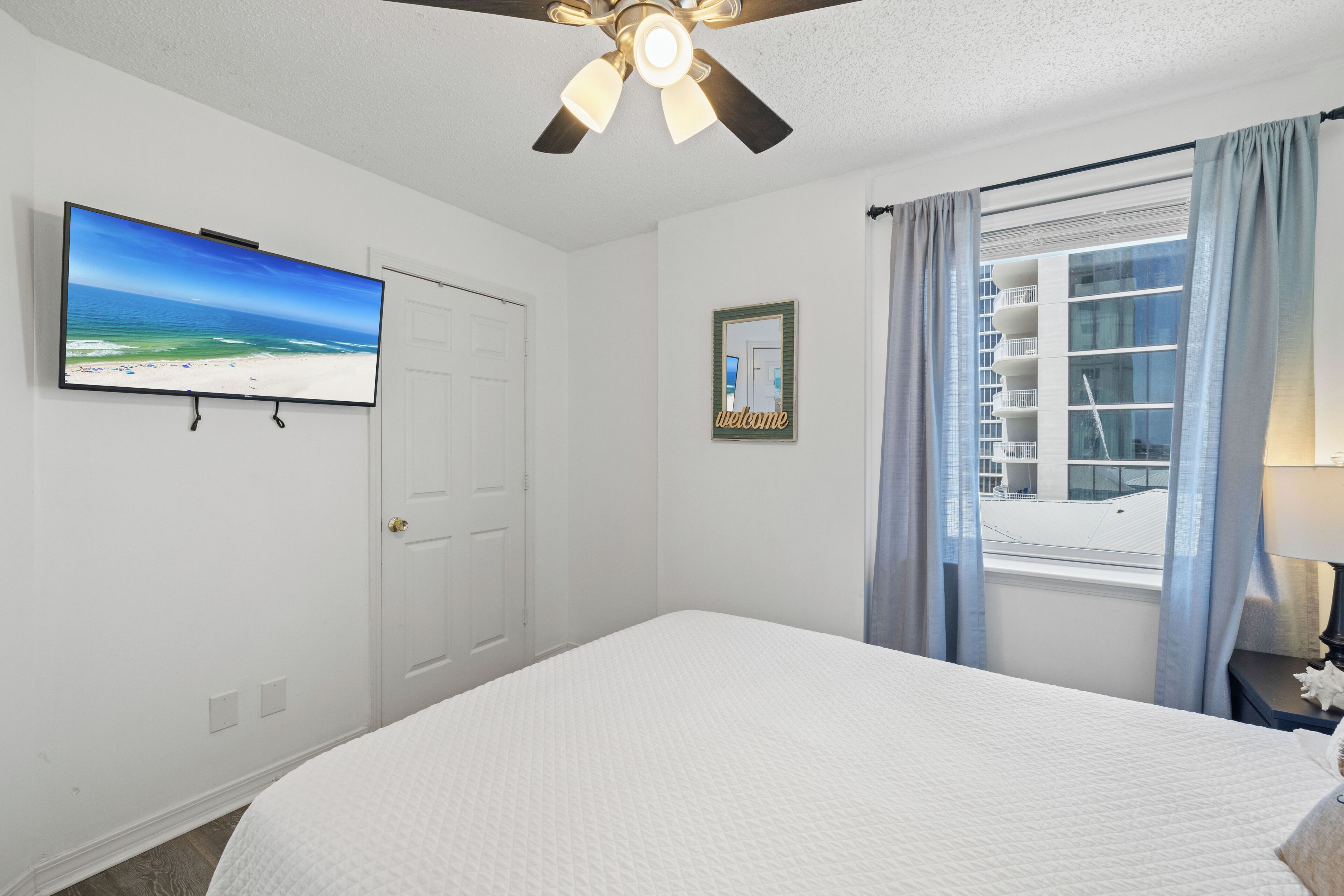 A hotel room with a bed, a ceiling fan, a TV on the wall, a window with curtains, and a welcome sign.
