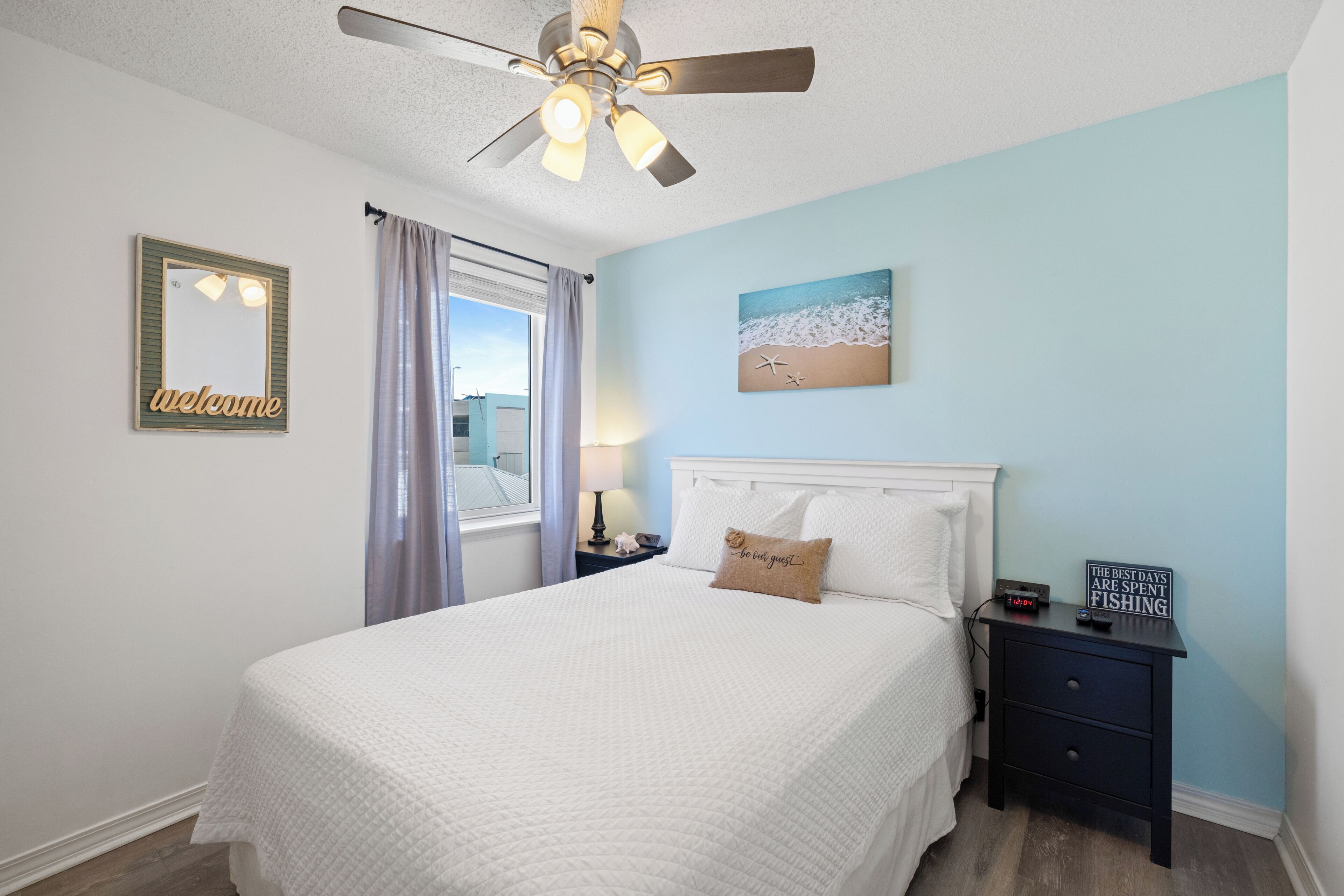 A bedroom with a bed, a nightstand, a wall-mounted clock, a ceiling fan, a window with curtains, and a welcome sign.