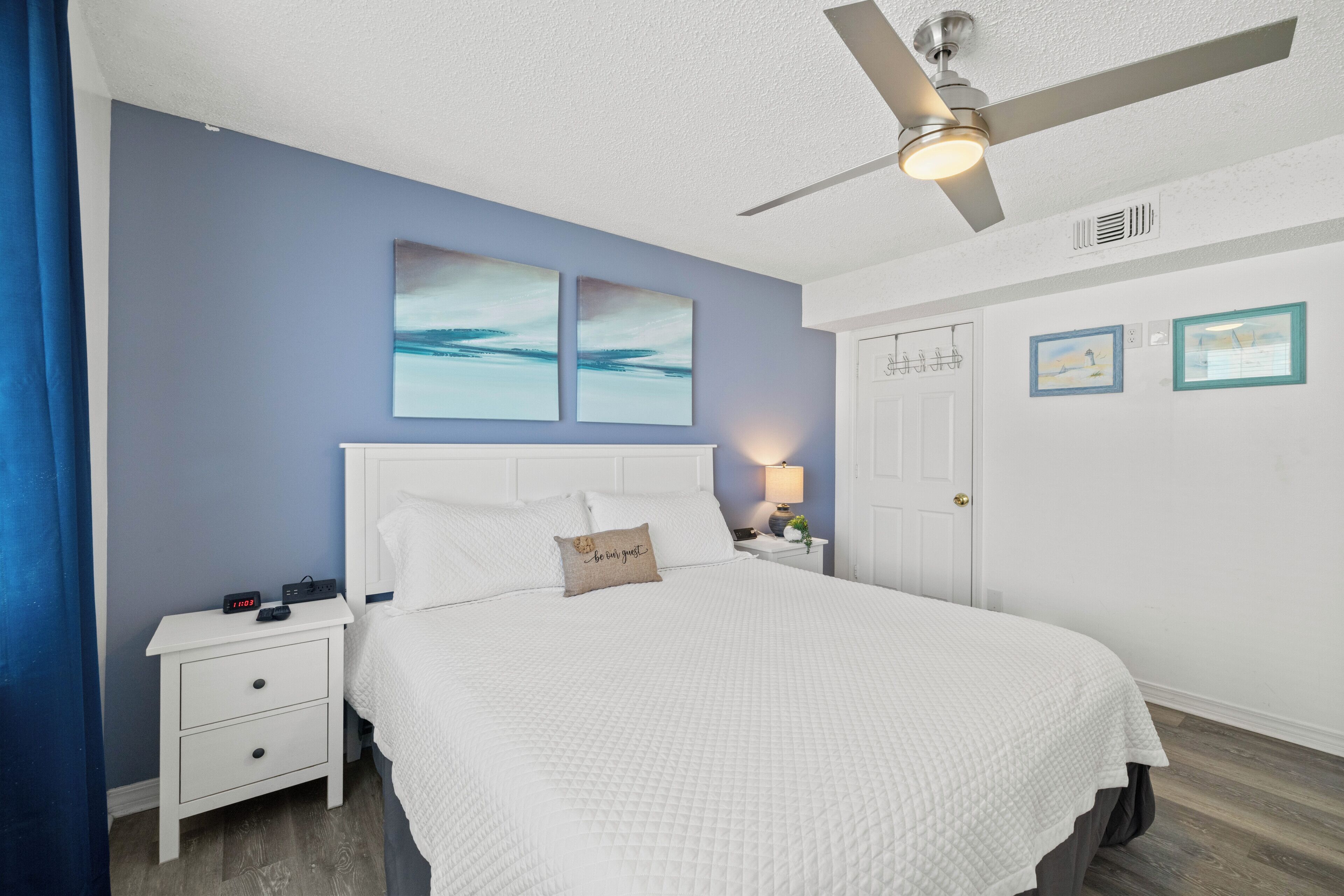 A bedroom with a bed, nightstands, a ceiling fan, artwork, and a door.