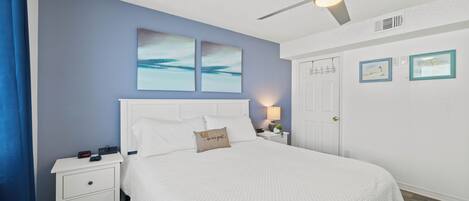 A bedroom with a bed, nightstands, a ceiling fan, artwork, and a door.