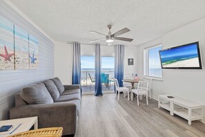 Ein Zimmer mit grauem Sofa, einem Esstisch mit Stühlen, einem Deckenventilator und Blick auf den Strand durch große Fenster.
