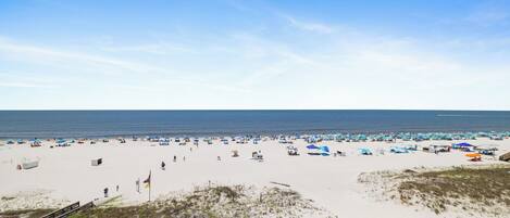 A beach with numerous umbrellas and beach chairs, a clear sky, and the ocean in the background.