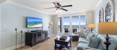 A spacious living room with a television, a ceiling fan, a balcony with a view of the beach, and a light blue color scheme.