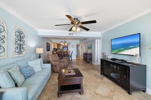 A spacious living room with a ceiling fan, a television displaying a beach scene, a dining area with a kitchen in the background, and a light blue wall. - Romar Tower 3B by Liquid Life (Orange Beach)