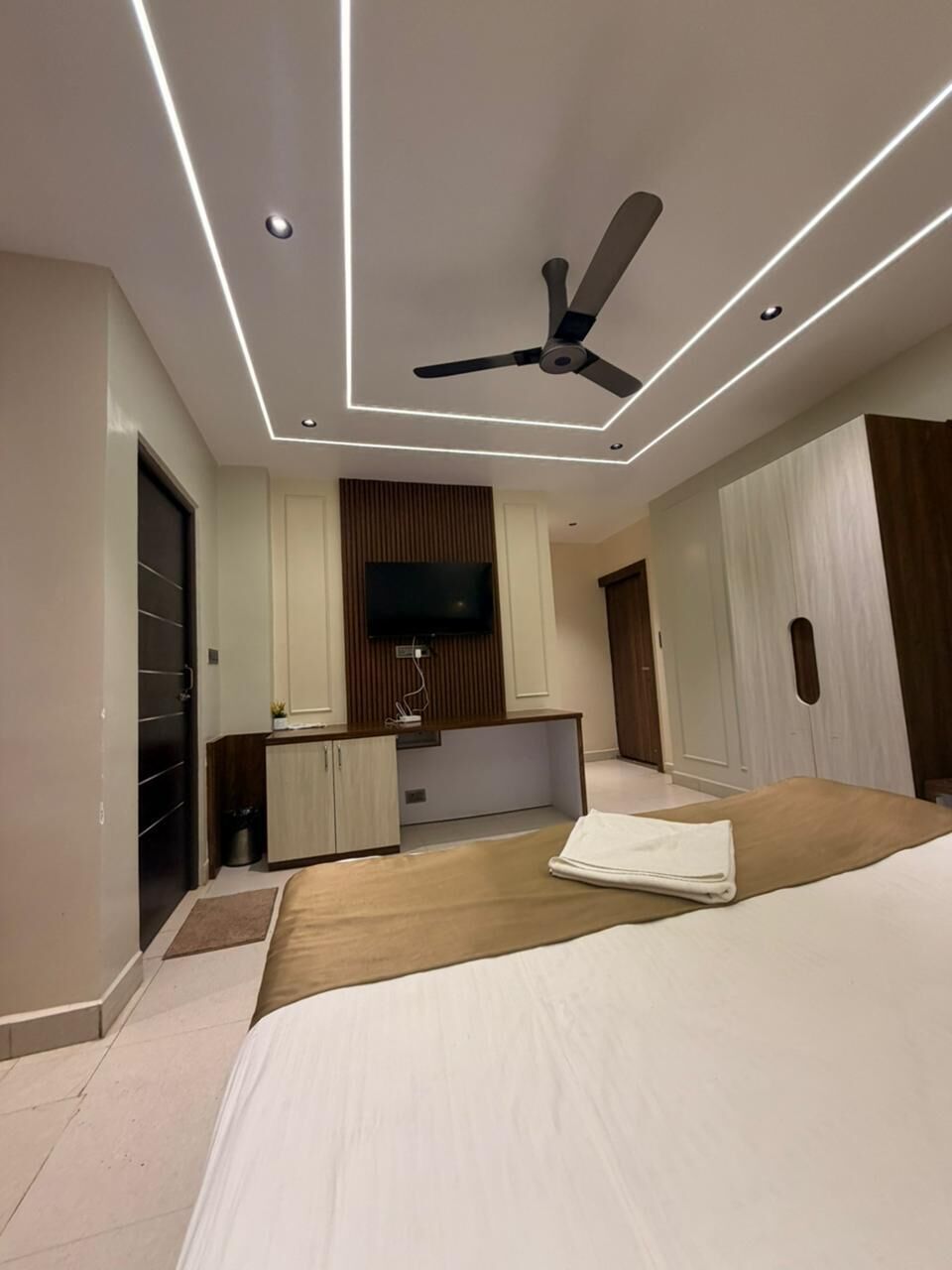 A modern hotel room with a bed, a ceiling fan, a TV, and a kitchenette.