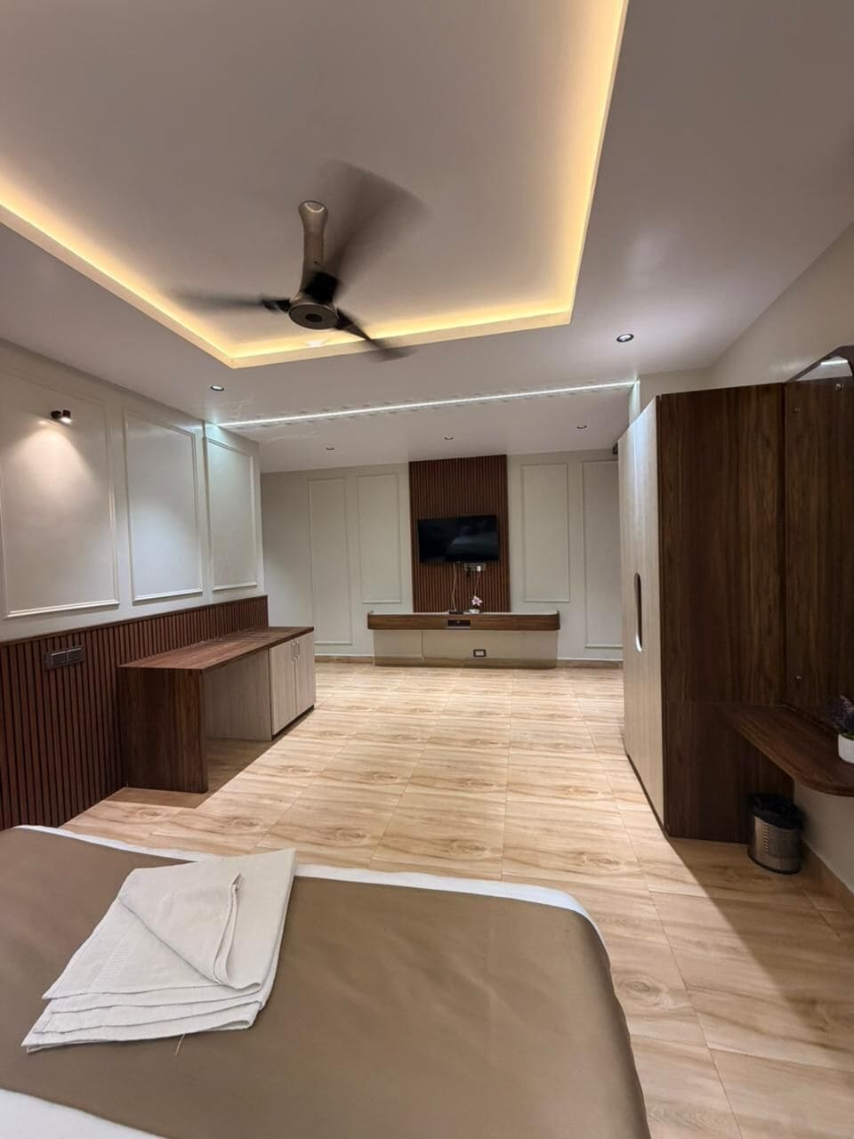 A modern hotel room with a wooden floor, a ceiling fan, a flat-screen TV, and a wooden cabinet.