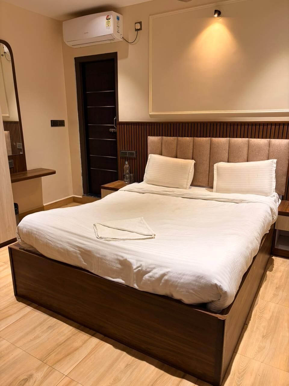 Una habitación de hotel moderna con un cabecero de madera, un espejo grande, un armario y un aire acondicionado instalado en la pared.