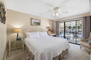 A hotel room with a bed, a nightstand, a lamp, a mirror, a desk with a chair, and a balcony with a view of the city. - Seaside Beach & Racquet 2203 by Liquid Life (Orange Beach)
