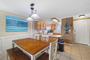 A modern kitchen with wooden cabinets, a dining table, and a large painting on the wall. - Seaside Beach & Racquet 2203 by Liquid Life (Orange Beach)