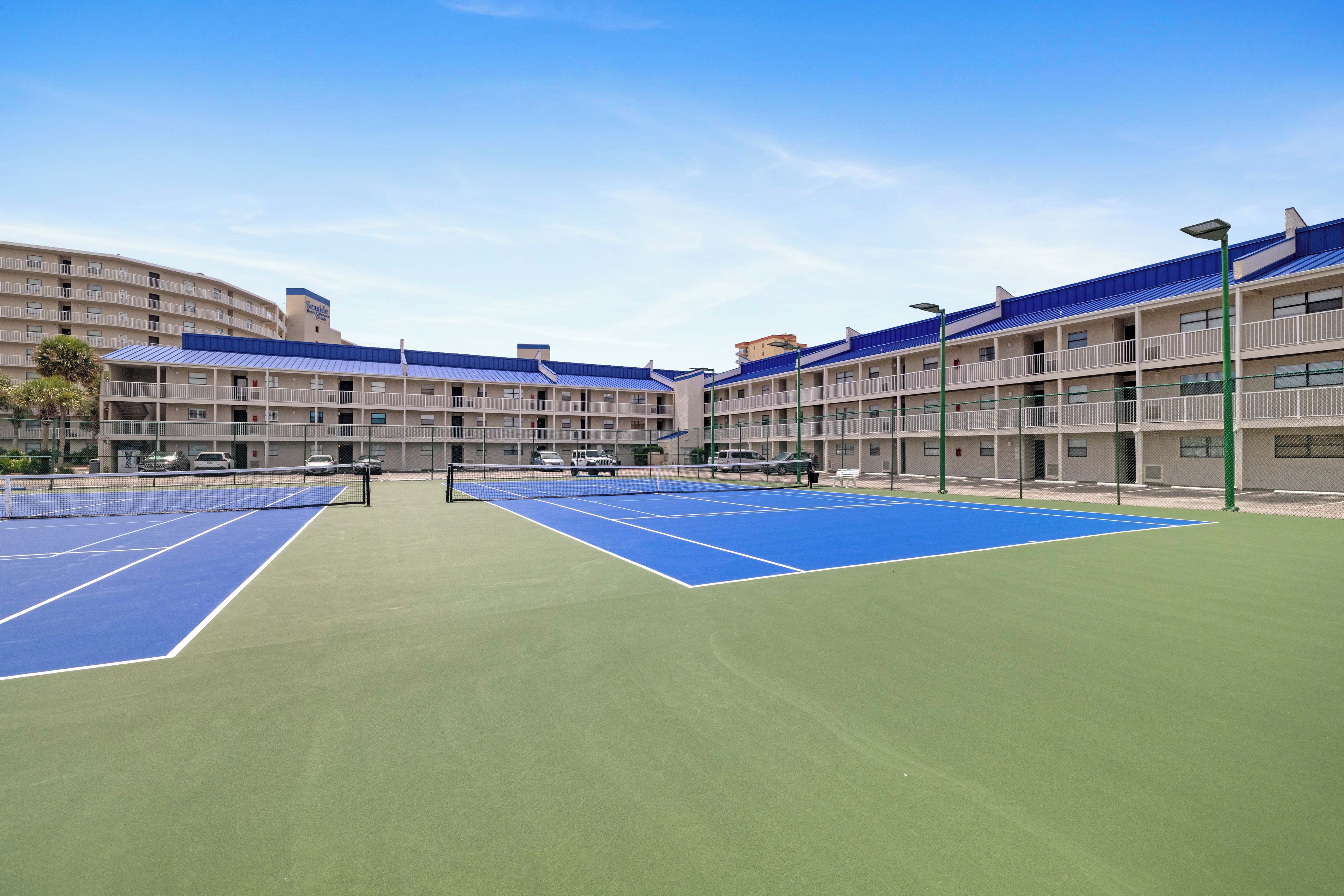 Un court de tennis avec un filet au milieu, entouré d’un bâtiment doté de balcons et d’un toit bleu.