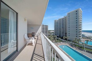 Een balkon met witte stoelen, een zwembad en uitzicht op een strand en hotelgebouwen.