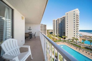 Een balkon met een witte stoel, een klein tafeltje en uitzicht op een zwembad en gebouwen.