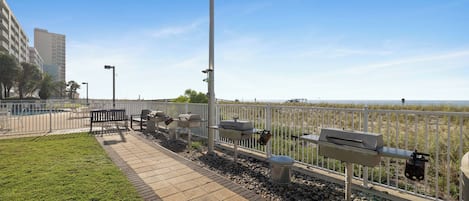 A paved walkway with benches and barbecue grills, bordered by a white fence, leading to a beach.