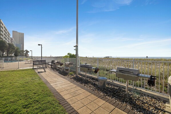 A paved walkway with benches and barbecue grills, bordered by a white fence, leading to a beach. - Surfside Shores 1803 by Liquid Life (Gulf Shores)