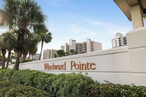 A sign with 'Windward Pointe' written on a white wall, surrounded by greenery and palm trees. - Windward Pointe 103 by Liquid Life (Orange Beach)