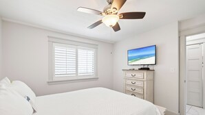 Un dormitorio con cama, ventilador de techo, televisor en una cómoda y una ventana con persianas.