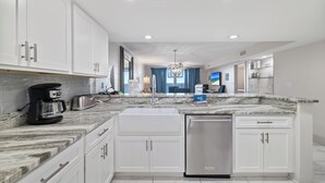 Cocina moderna con gabinetes blancos, encimera de mármol, lavavajillas integrado y vista al área de estar.