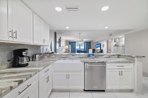 A modern kitchen with white cabinets, a marble countertop, a built-in dishwasher, and a view to the living area. - Wind Drift 611SW by Liquid Life (Orange Beach)
