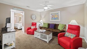 Un salon avec un ventilateur de plafond, un canapé, deux fauteuils, une table basse, un téléviseur et une lampe.