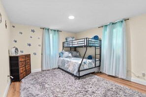 Een stapelbed met een bedspreider met patroon, een dressoir en een zacht vloerkleed in een kamer met lichte muren en gordijnen.