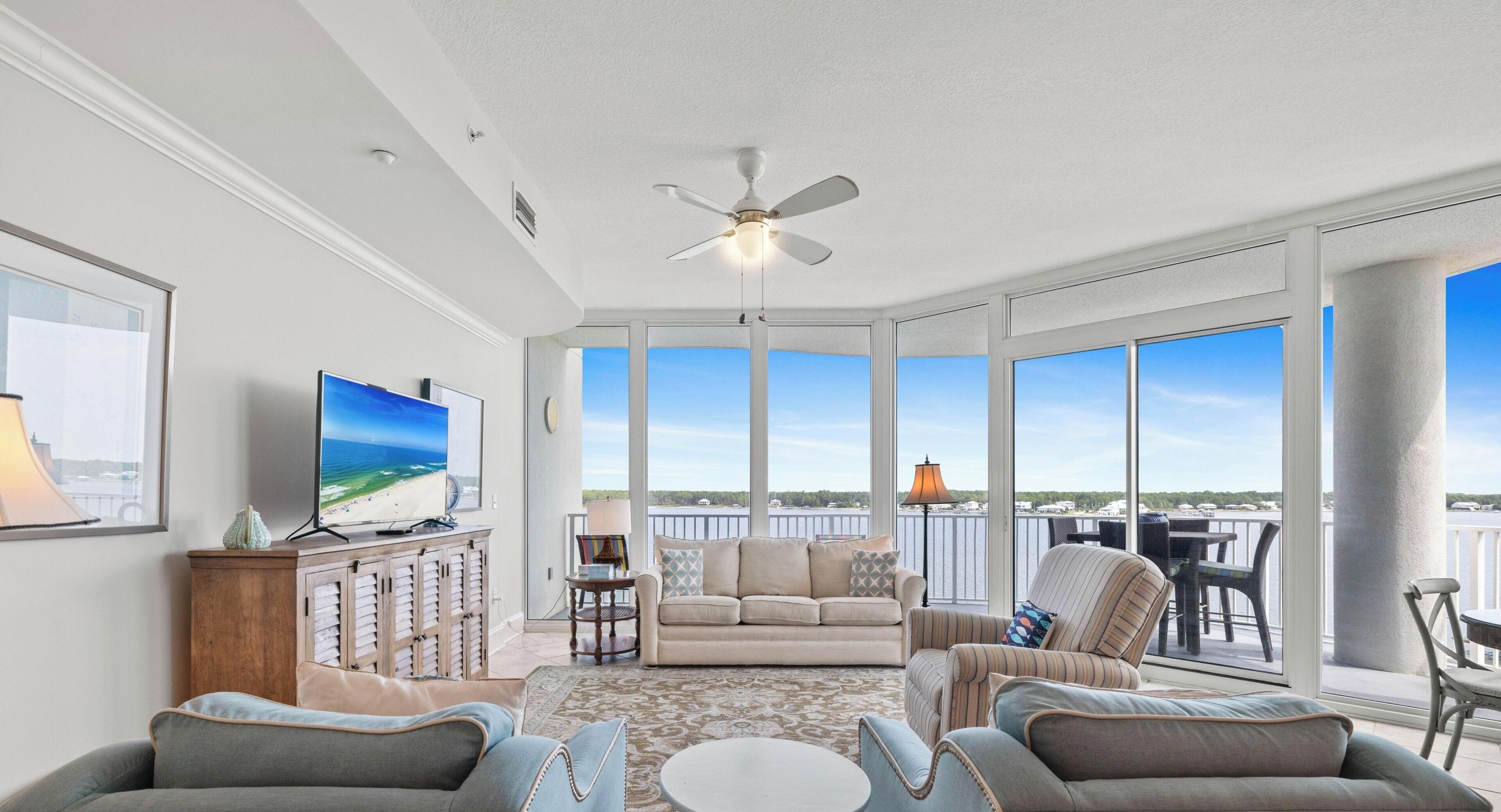 A spacious living room with a large TV, comfortable seating, and a view of a lake through large windows.