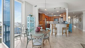 A modern kitchen with wooden cabinets, a central island, and a dining area with glass tables and metal chairs. - Bel Sole 603 by Liquid Life (Gulf Shores)