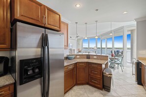 A modern kitchen with wooden cabinets, a stainless steel refrigerator, and a large island with a sink. - Bel Sole 603 by Liquid Life (Gulf Shores)